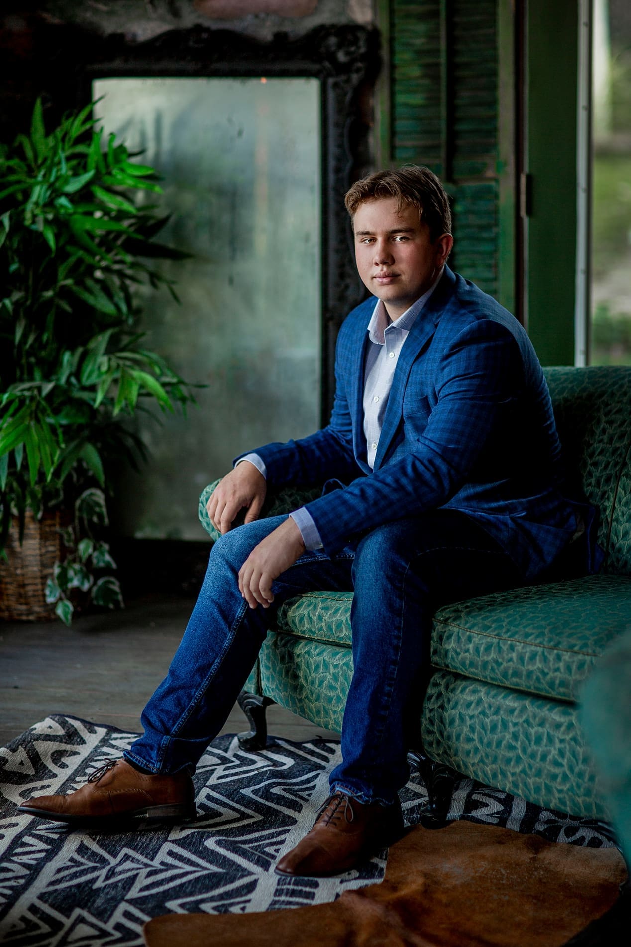 Indianapolis senior guy portrait in a blue blazer and jeans sitting on a vintage green sofa inside a stylish studio during a senior picture session.