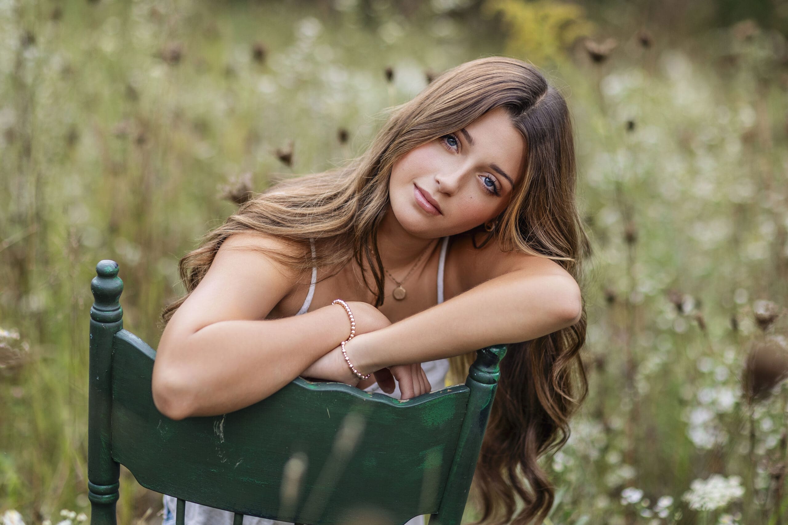 Indianapolis senior girl with long brunette hair leaning on a vintage green chair in a wildflower field during a natural outdoor senior portrait session.
