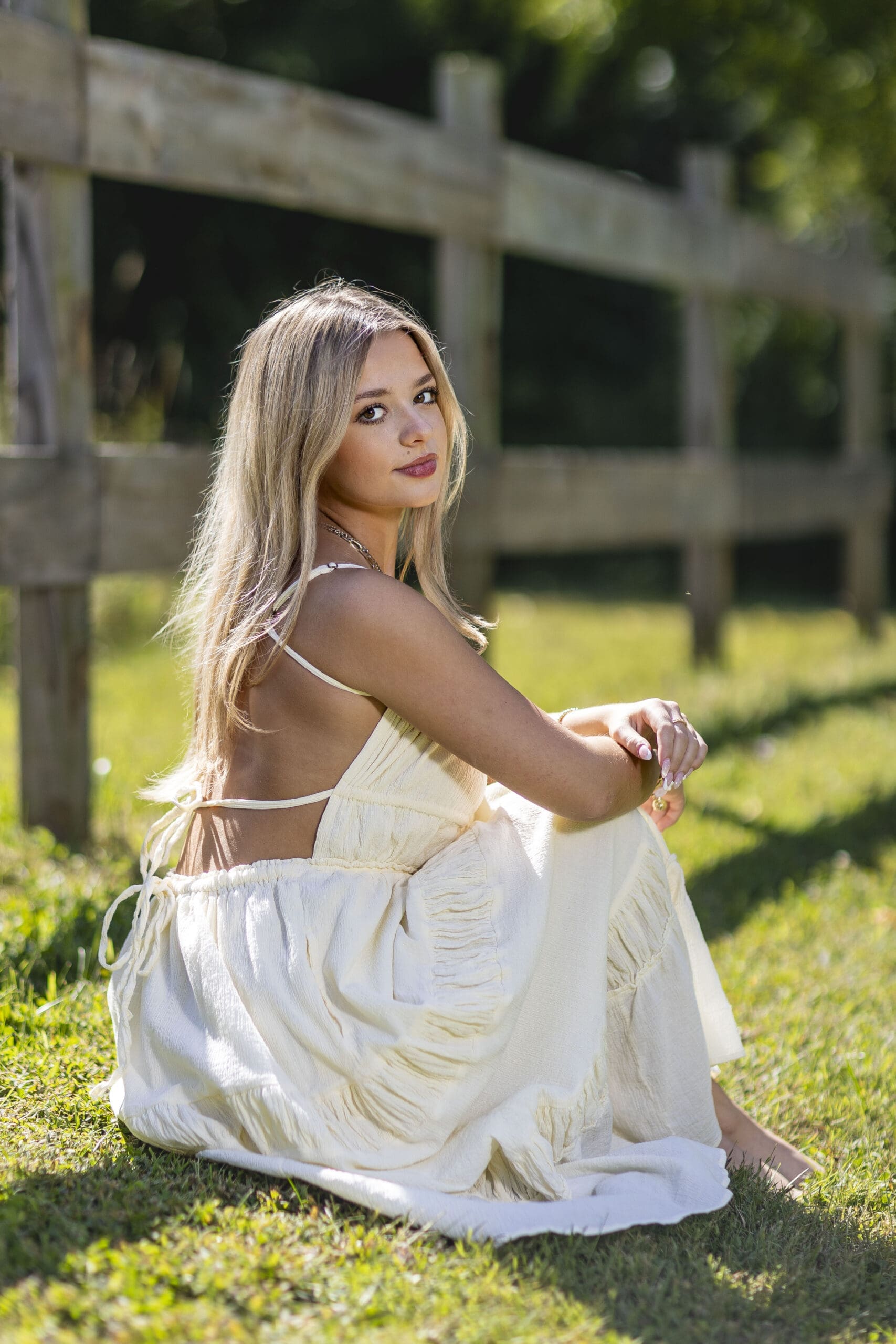 Indianapolis senior girl in a flowing cream dress sitting on a rustic wooden fence surrounded by soft greenery and golden sunlight during an outdoor senior picture session.