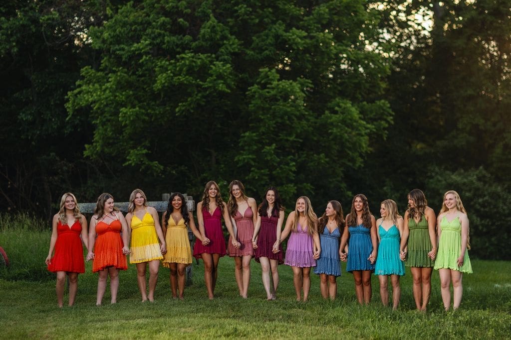 MAC Senior Model Team members standing hand in hand in a grassy field at sunset, wearing colorful dresses in a rainbow of colors during a group senior photography session in Indianapolis
