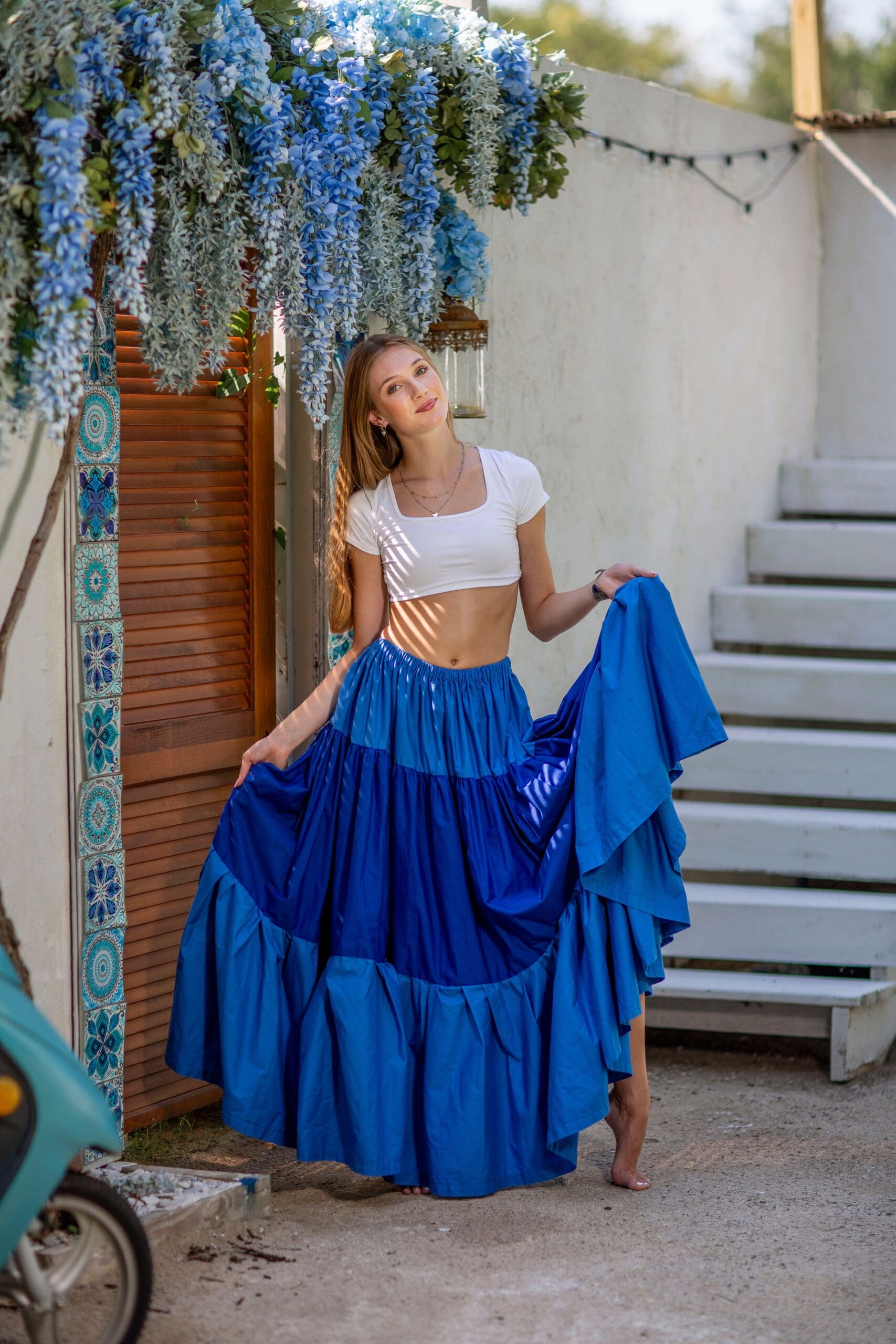 High school senior girl posing barefoot in a flowing royal blue skirt and white crop top beneath cascading blue flowers at the Marci & Christy Studio Backlot in Indiana.