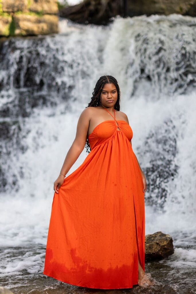 High school senior standing barefoot in a flowing orange dress in front of a waterfall during an Indiana senior picture session, creating a bold and dramatic portrait.